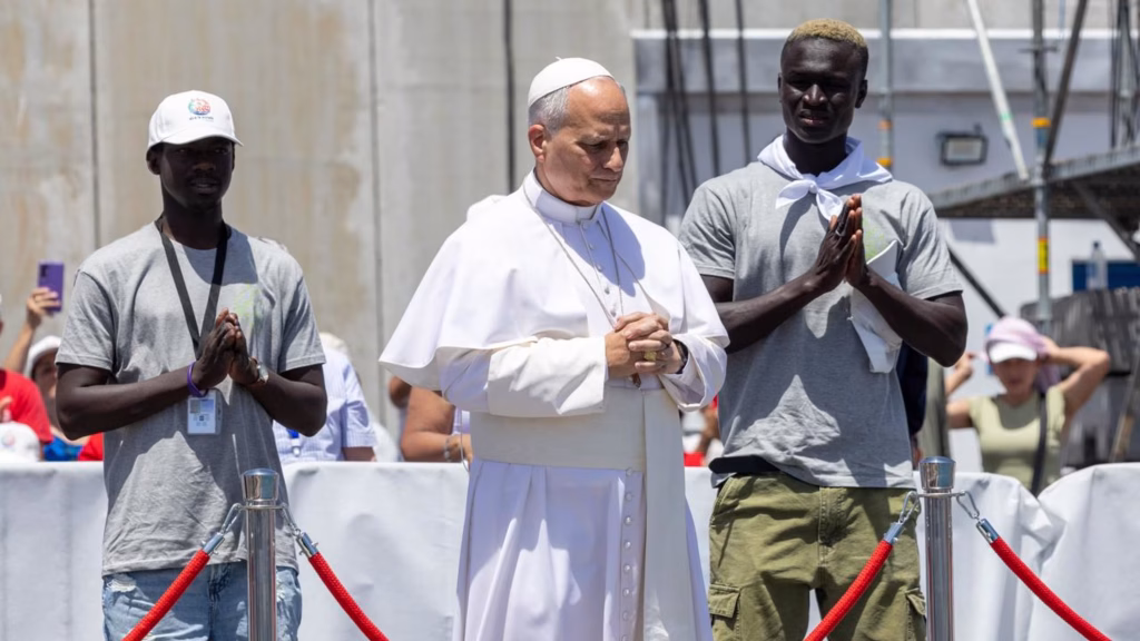 Pope and two migrants stand behind roped area on small fishing dock praying hands clasped.