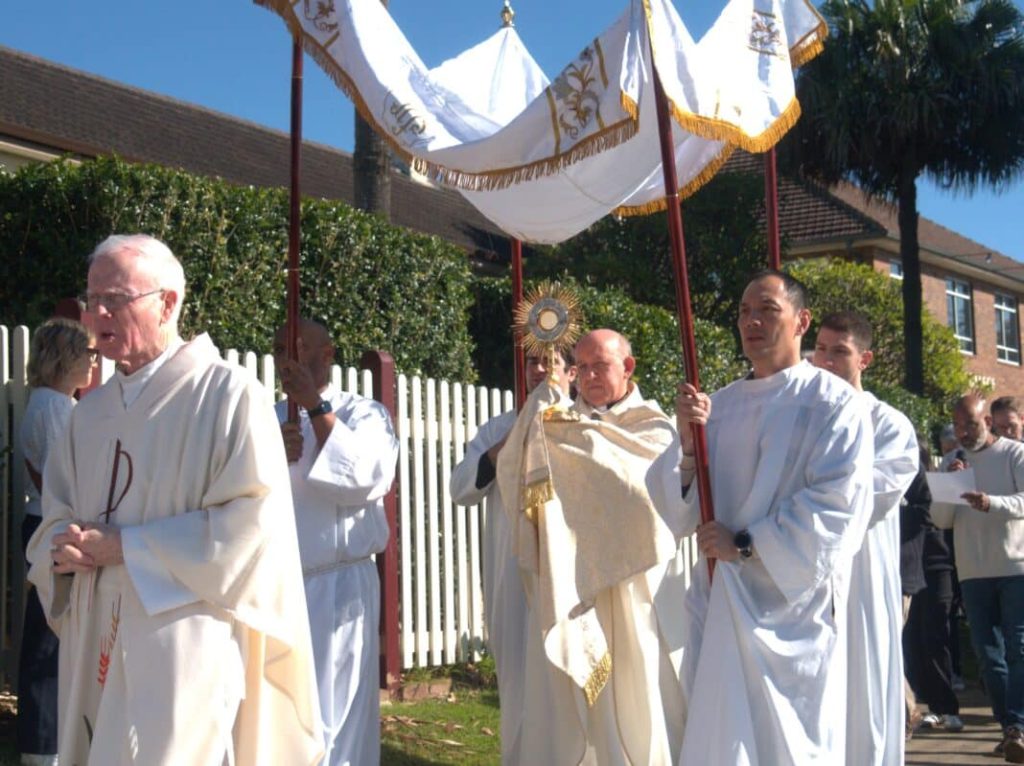 Priest holding Eucharist under canopy being held by 4 servers in a Corpus Christi procession on an outdoor sunny street.